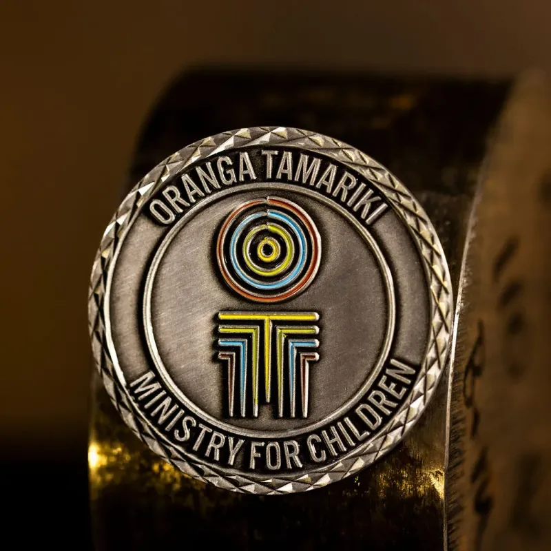 Custom New Zealand Ministry For Children/Oranga Tamariki challenge coin in antique silver finish, with the ministry's logo in the centre. The logo is coloured with enamel. The edge is cross-cut.
