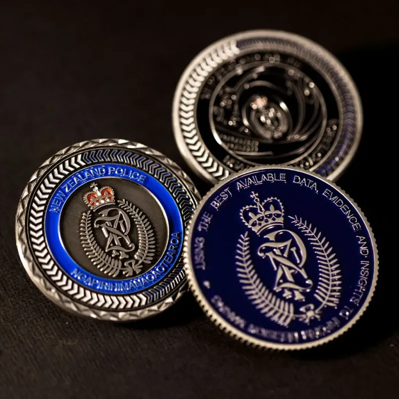 Three custom New Zealand Police challenge coins in antique silver finish, with the New Zealand police logo in the centre of each. The logo is coloured in with enamel. One coin has dark blue enamel, one has black enamel, the other has light blue enamel. The challenge coins features cross-cut edges.